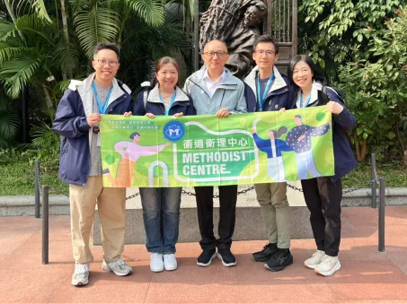 循道衛理中心職員於戶外雕塑前舉起中心橫幅合影